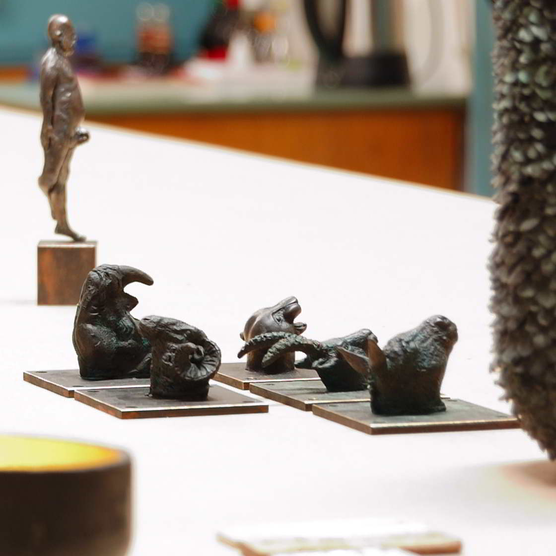 kleine Tierkopf Skulturen wie Widder, Nashorn, Esel, Steinbock und Jaguar. Im Hintergrund ist ein männlicher Akt in Bronze gegossen und auf einem Kupfersockel stehend zu sehen. Im Vordergrund ist ein Gefäß angeschnitten, dass eine sehr meditative Aura hat.